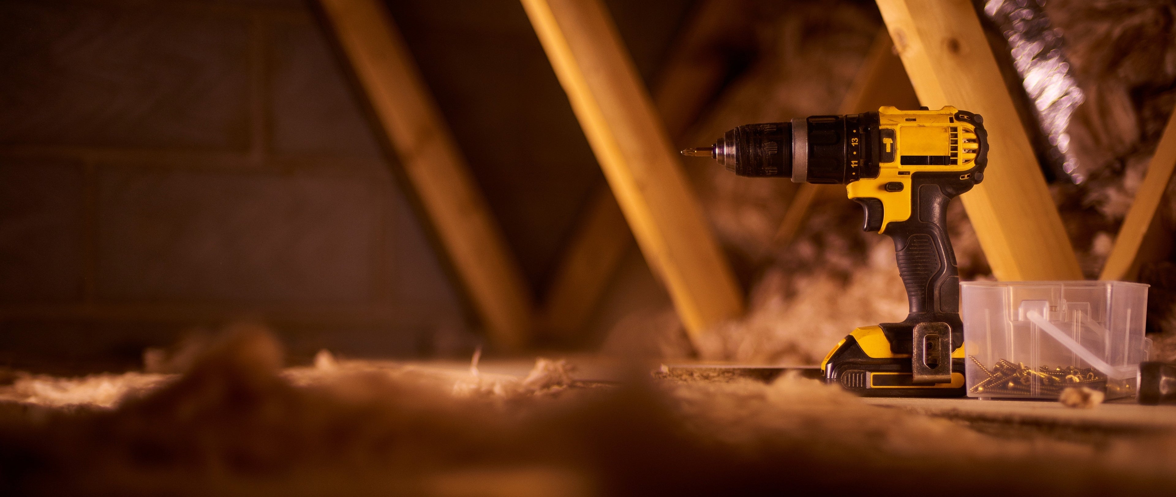 A yellow and black cordless drill resting on a dusty workbench next to a small clear container of screws, with wooden beams and insulation visible in the background.