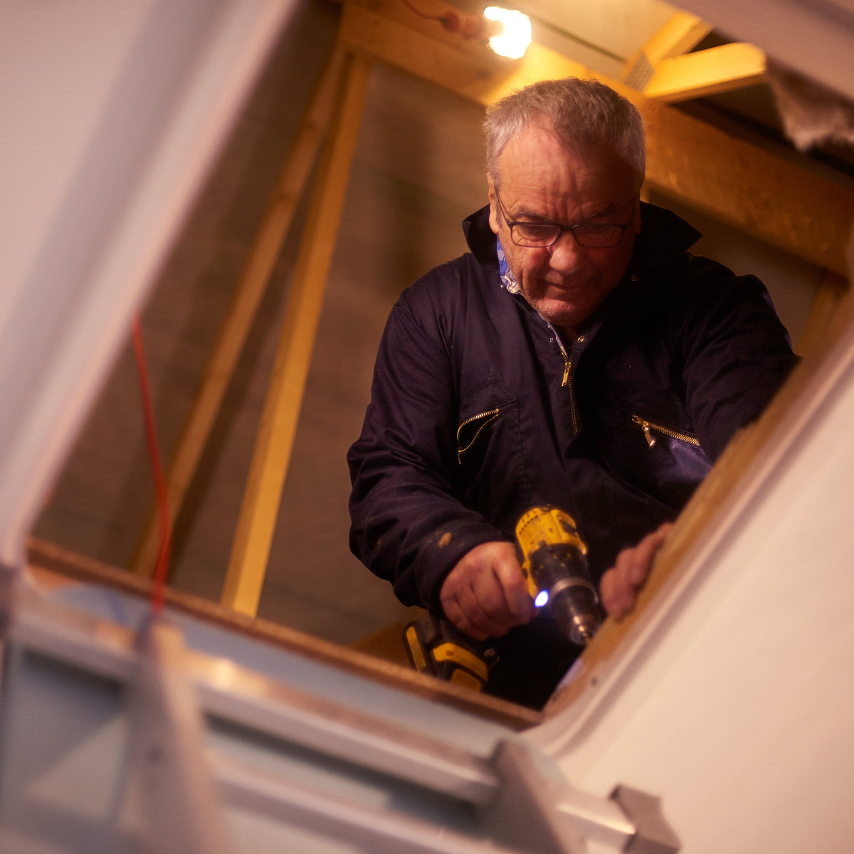 A man wearing safety glasses and a dark work jacket uses a yellow cordless drill inside a narrow, unfinished attic or loft space with wooden beams and a light fixture overhead.