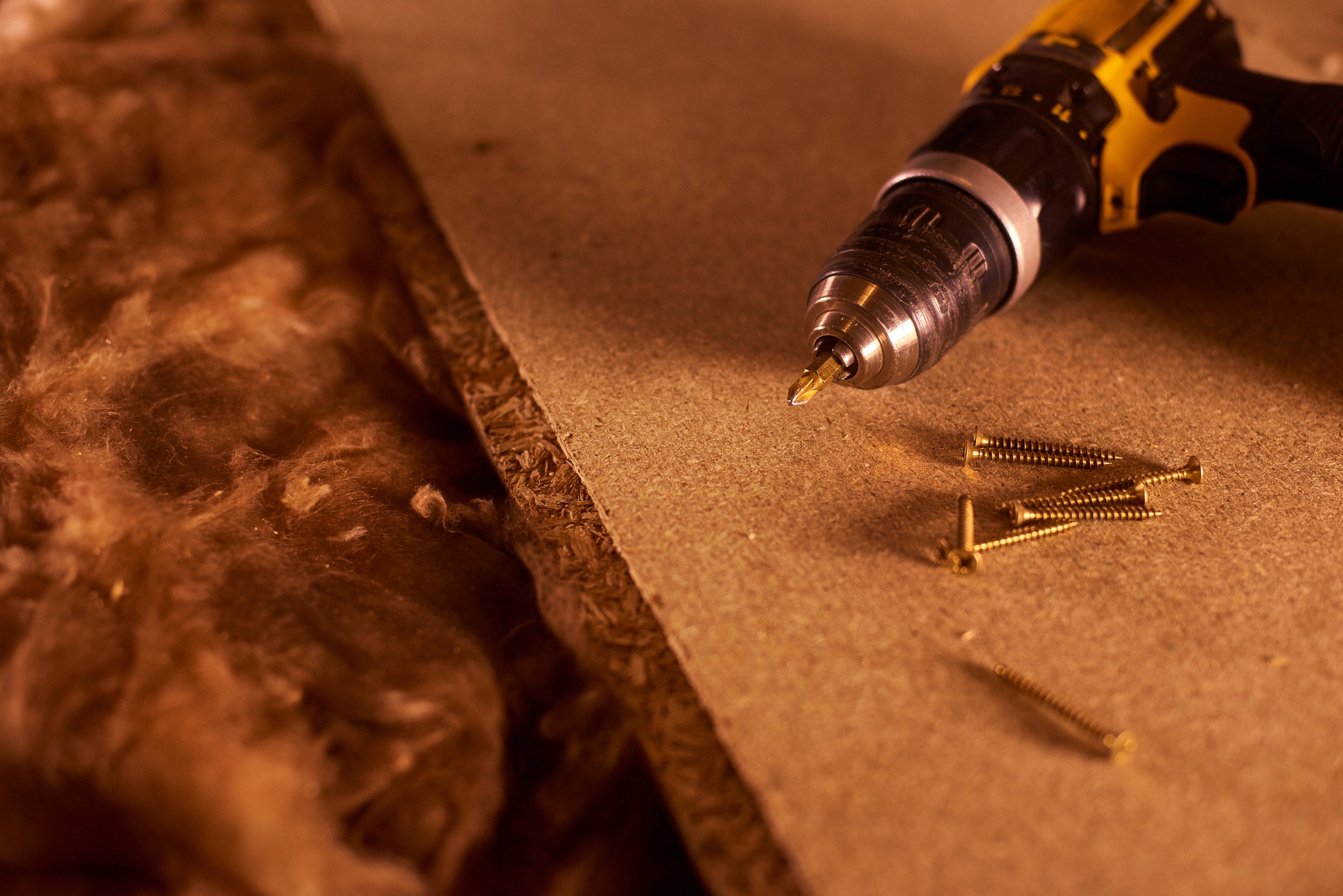 A yellow and black cordless drill with a metal bit hovering over a brown particle board sheet, with several loose gold-colored screws scattered nearby on a warm, dimly lit surface.